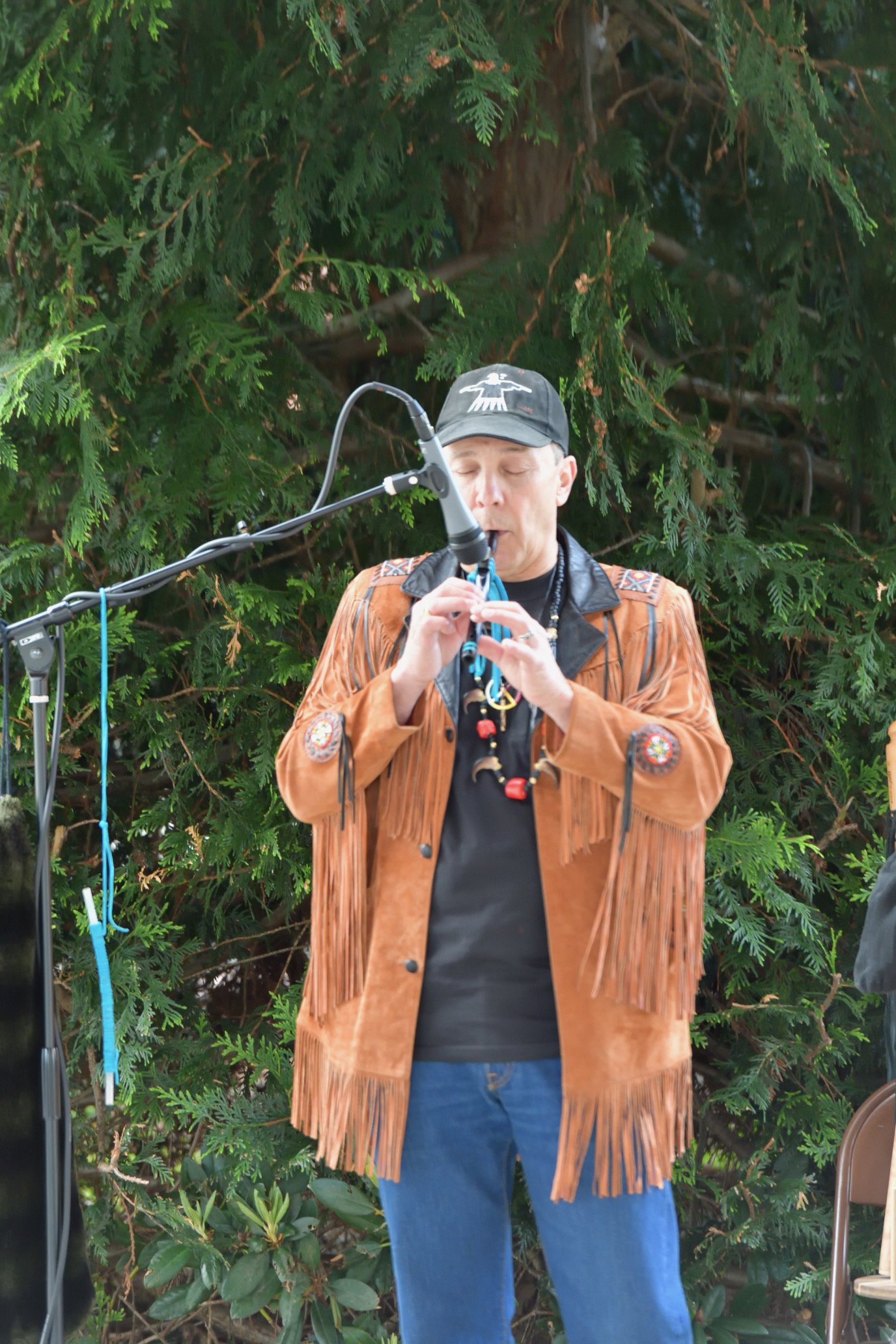 Man in a fringed leather jacket and black cap playing a wind instrument into a microphone outdoors