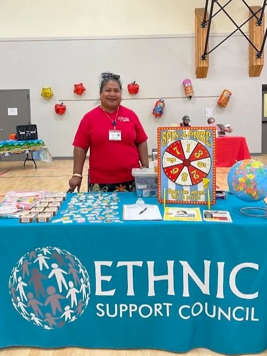 Volunteer at an Ethnic Support Council booth with a colorful spin-to-win prize wheel and informational materials at a community event.