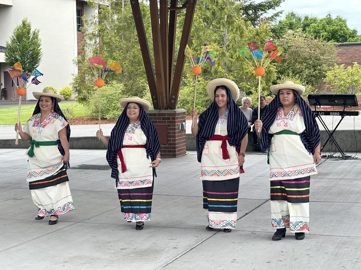Four women in traditional embroidered dresses and sombreros perform with colorful flags outdoors