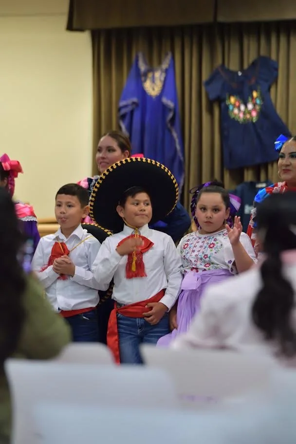 Children in traditional Mexican costumes pose indoors, including a boy wearing a large sombrero and girls in colorful folkloric dresses.