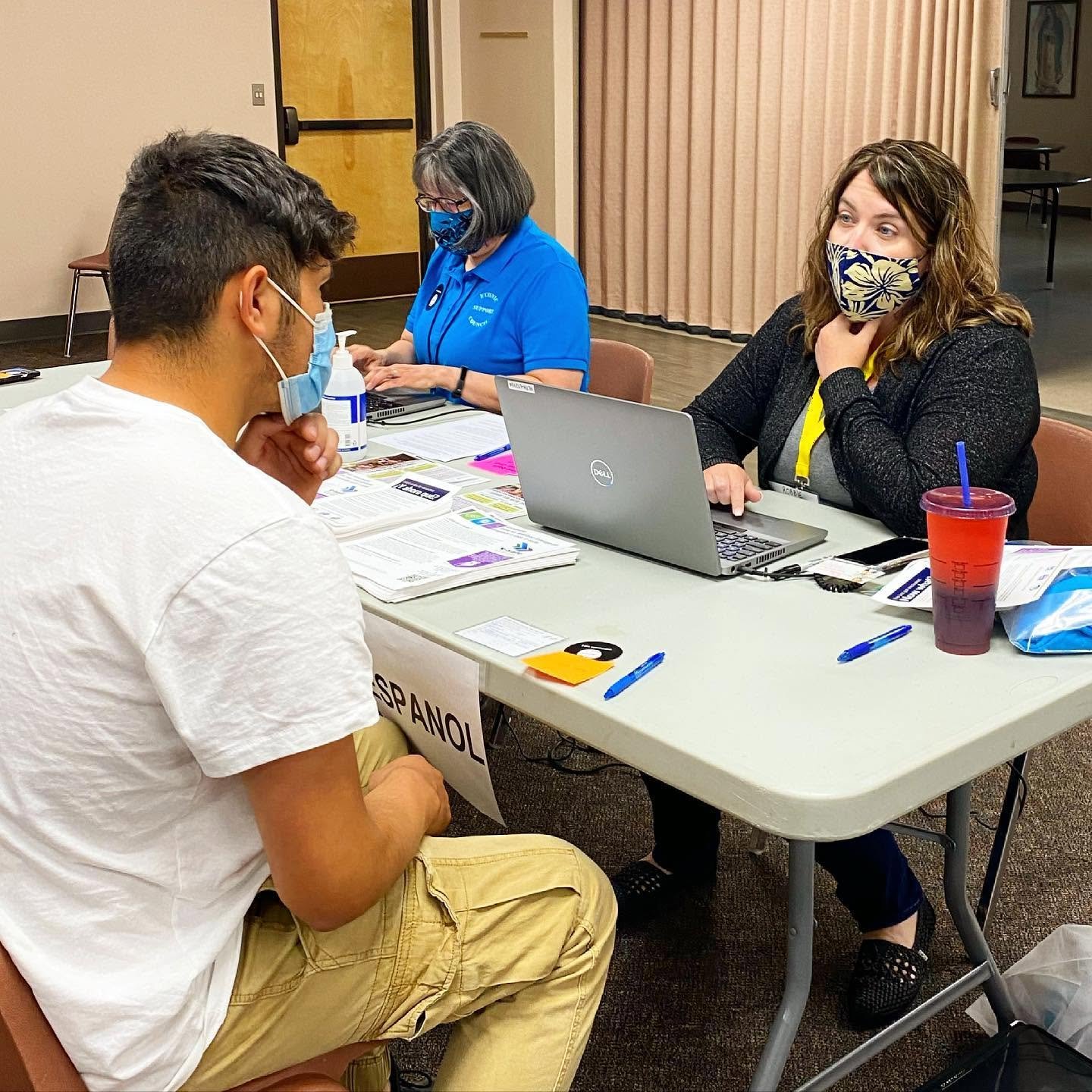 Masked man speaking with two masked staff members at an information table with laptops and brochures