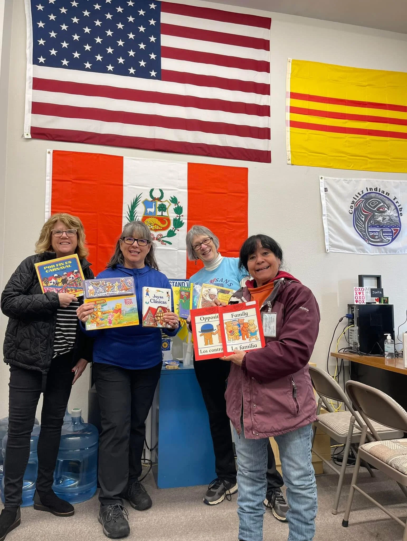 Four women hold children’s books in front of American, Peruvian, and other cultural flags at a community event.