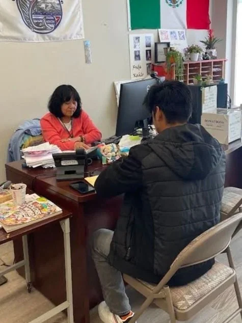Two people meeting at an office desk, with a Mexican flag, plants, paperwork, and computer monitor in the background.