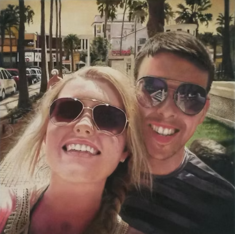 Two young adults wearing sunglasses are smiling and taking a selfie outdoors with palm trees and buildings in the background.