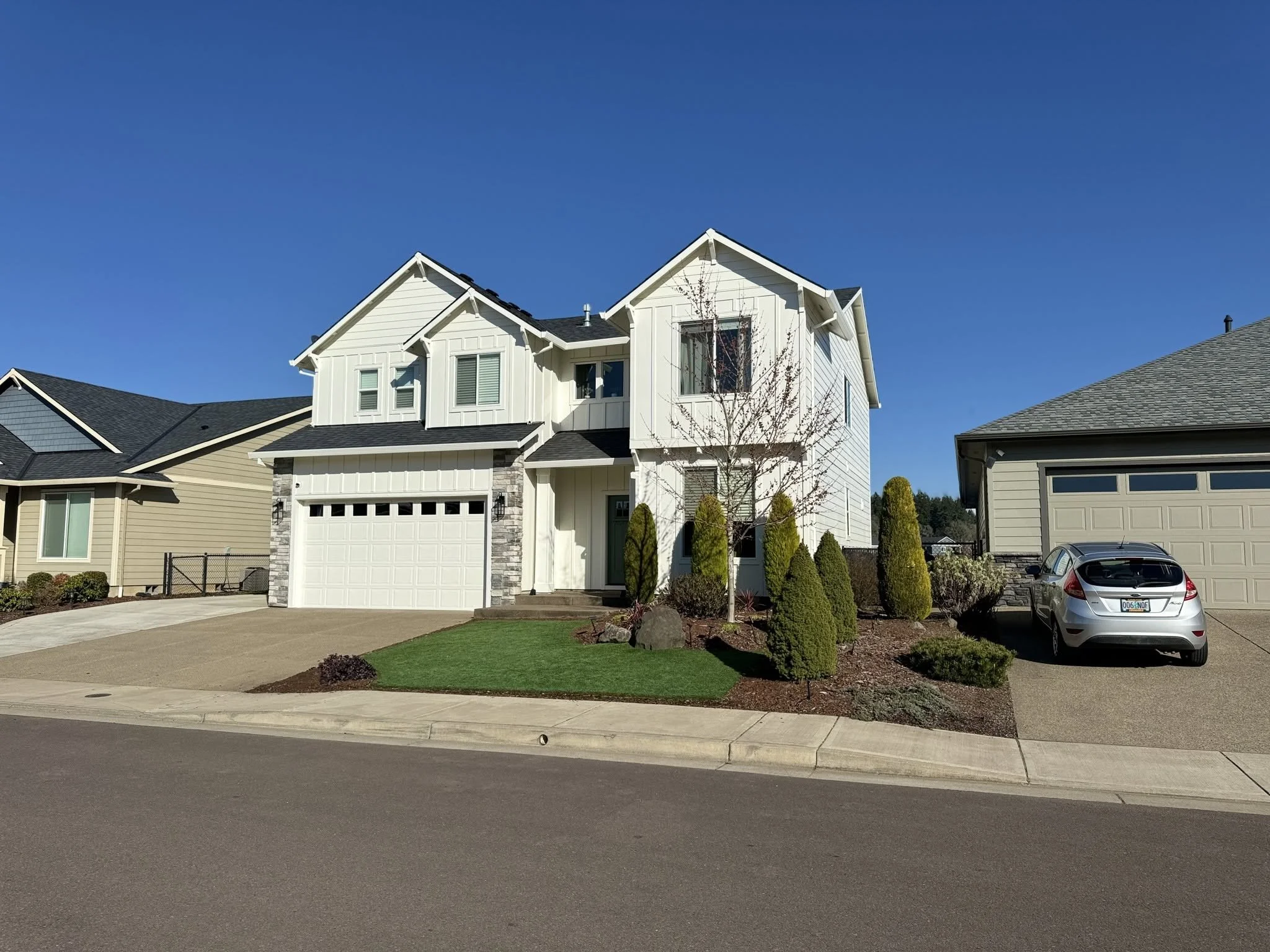 A modern two-story house with white exterior siding, black roof, and a two-car garage. There are small trees, shrubs, and a lawn in the front yard. A gray car is parked in the driveway, and neighboring houses are partially visible on either side. The