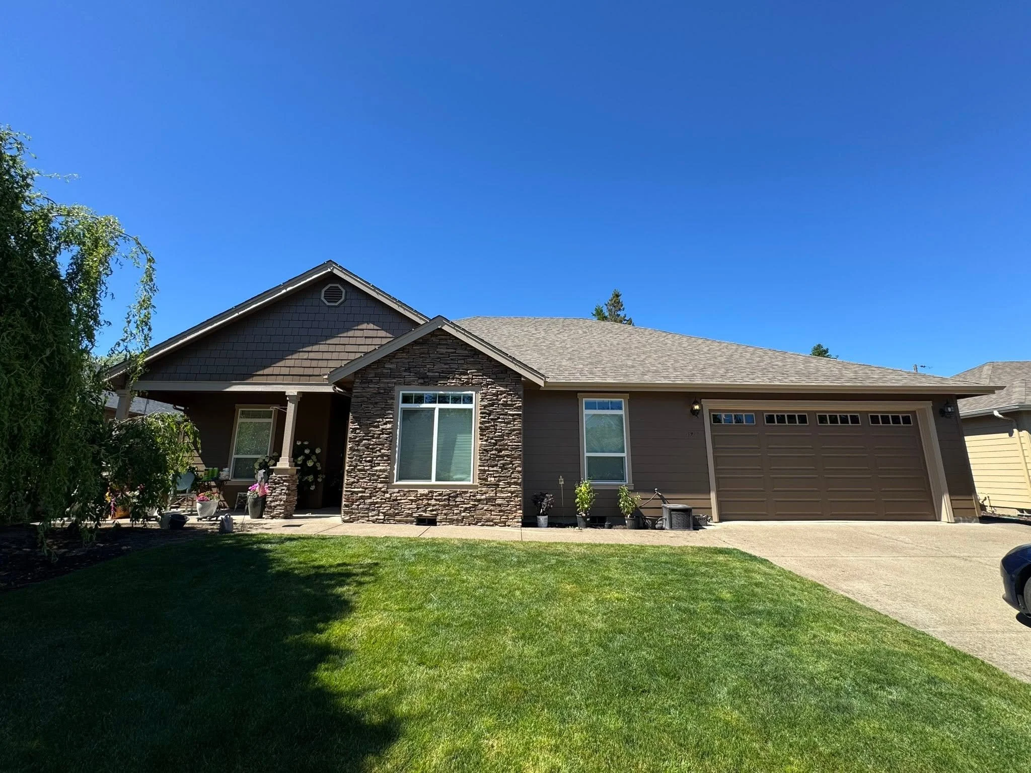 Front view of a suburban house with gray siding, stone accents, a two-car garage, and a well-kept lawn under a clear blue sky.