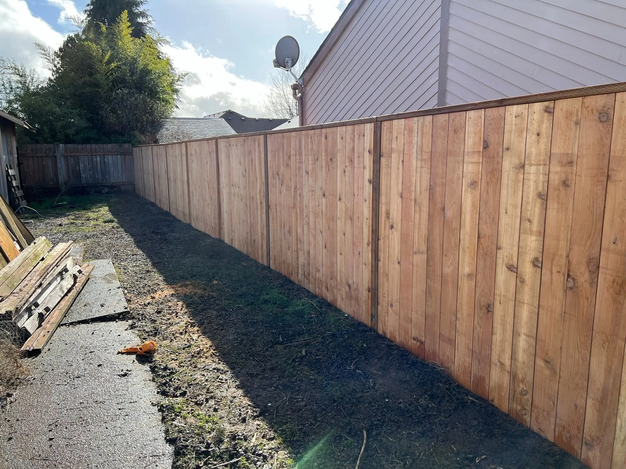 A backyard with a wooden fence, a satellite dish on the side of a house, and a cloudy sky.