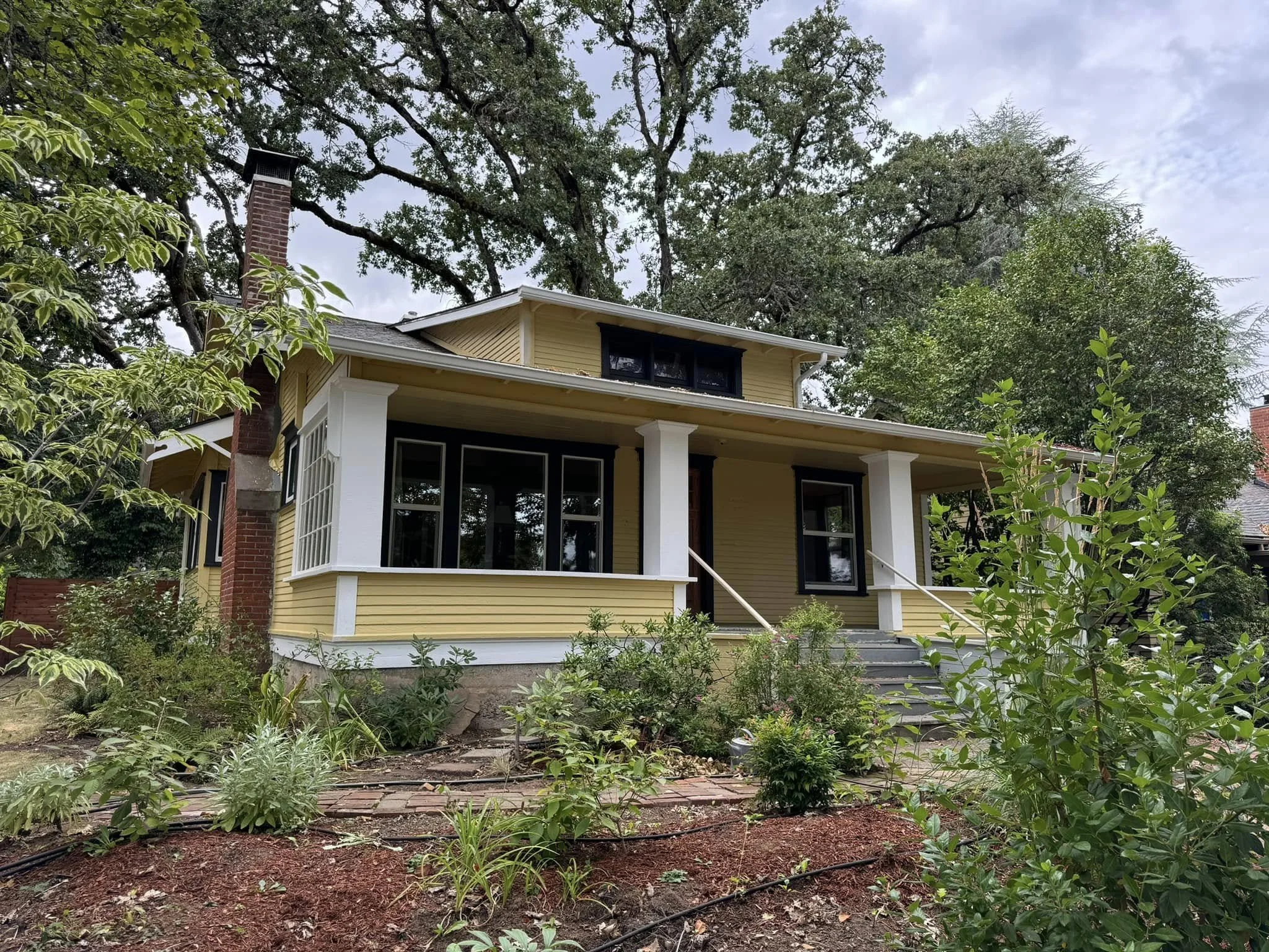 A yellow house with white columns and black window shutters surrounded by green trees and plants, with a cloudy sky overhead.