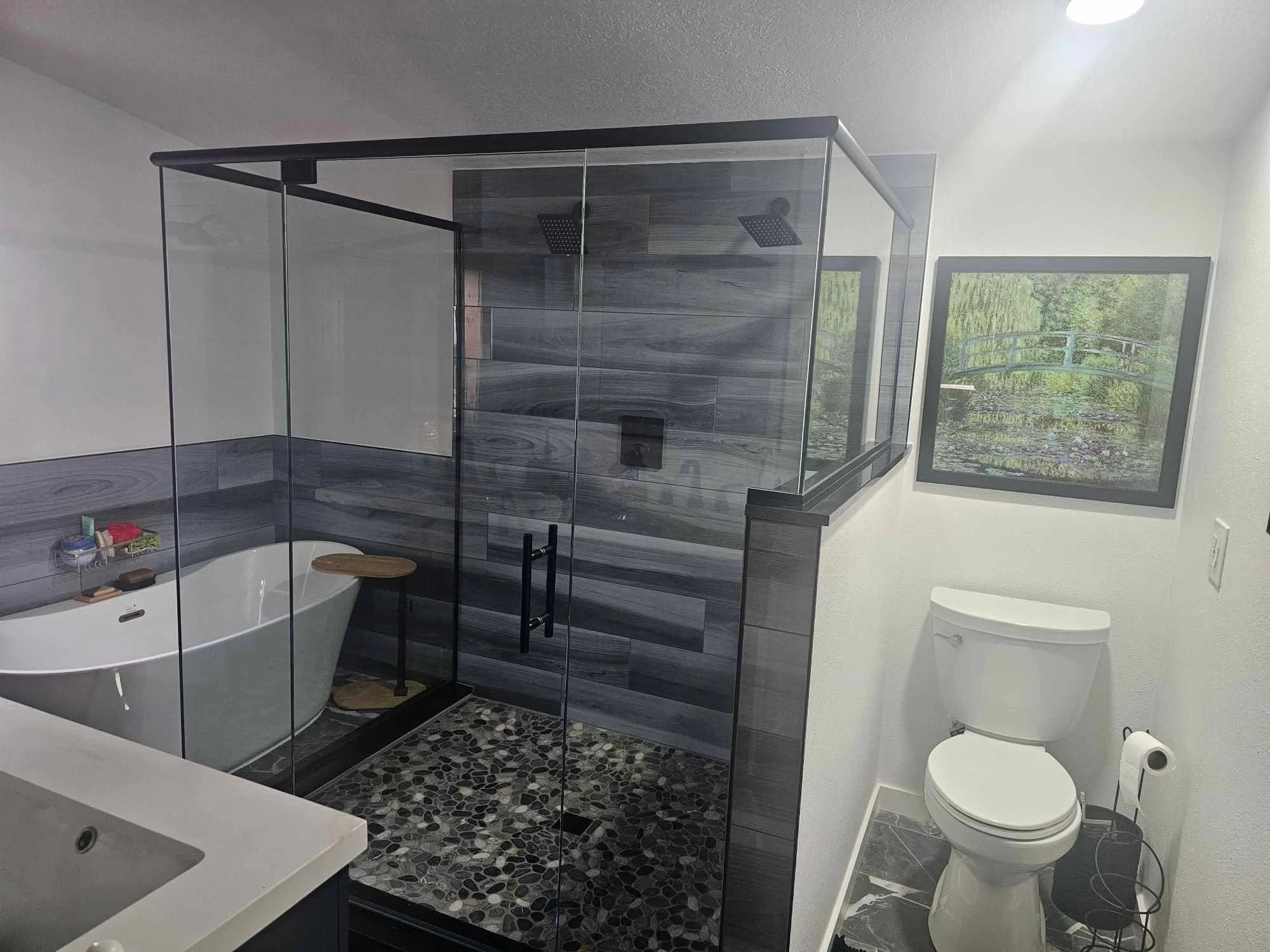 Modern bathroom with a glass-enclosed shower, a bathtub with a wooden tray, a toilet, a large window showing greenery outside, and a wall-mounted cabinet.