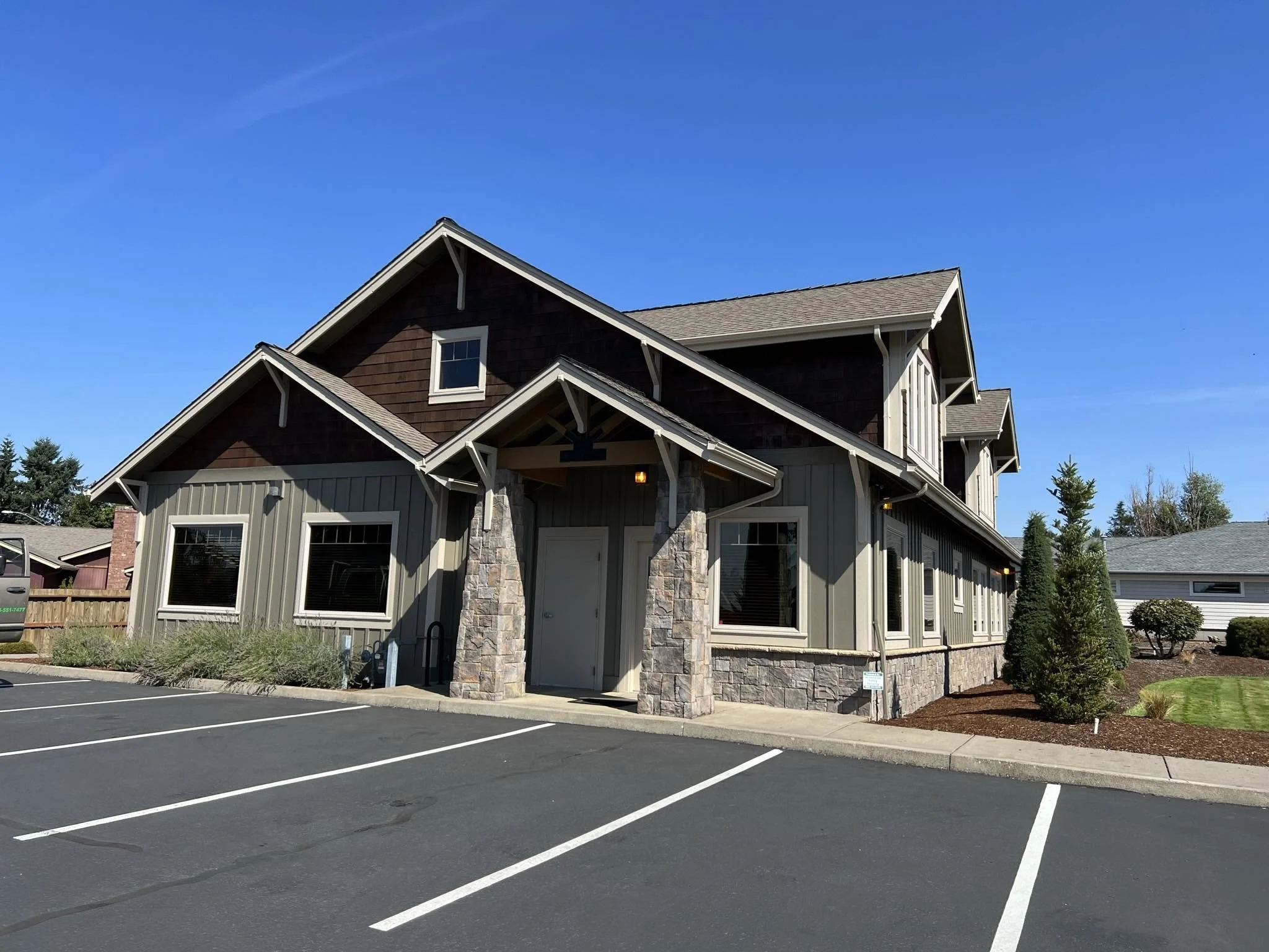 A modern house with brown siding, stone accents, and a gable roof, surrounded by a parking lot with marked parking spaces and landscaped bushes under a clear blue sky.