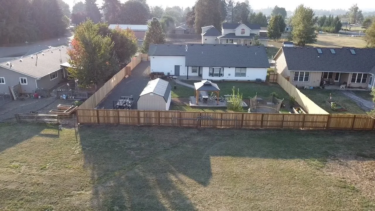 View of a backyard with a shed, a small pavilion, and a trampoline, enclosed by a wooden fence, with neighboring houses and trees in the background.