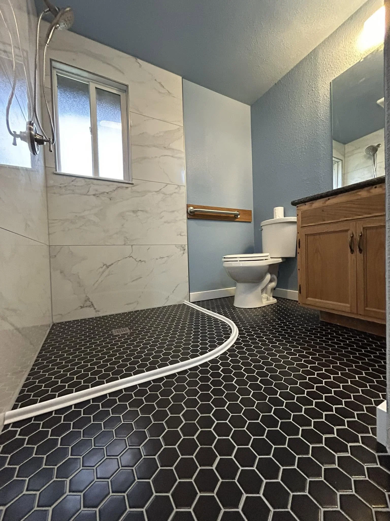 A bathroom with black hexagonal floor tiles, a wooden vanity with a granite top, a toilet, a small window, and a shower area with a grab bar and a hose.