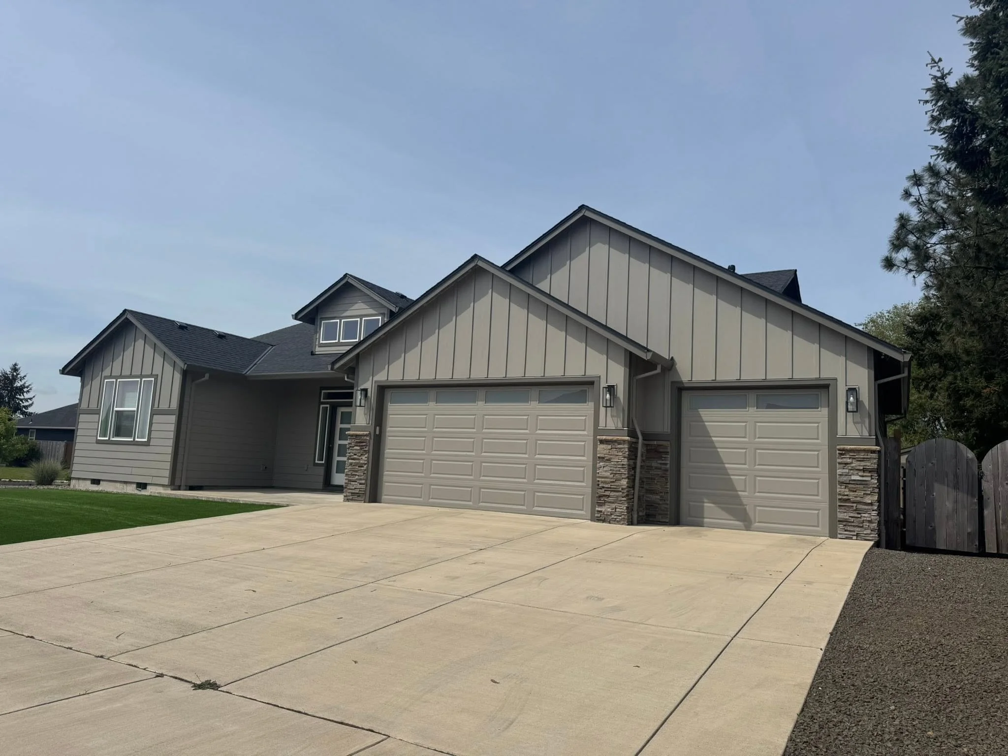 A two-story house with gray vertical siding, white trim, and a gable roof with black shingles. There is a two-car garage with beige doors, a concrete driveway, and a front yard with grass and plants. A tall tree is on the right side of the house.
