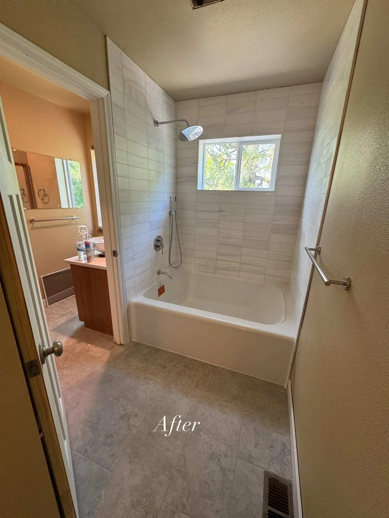 A bathroom after renovation showing a bathtub with a shower head, a window, a towel bar, and a doorway leading to a vanity area with a mirror, sink, and water bottle.