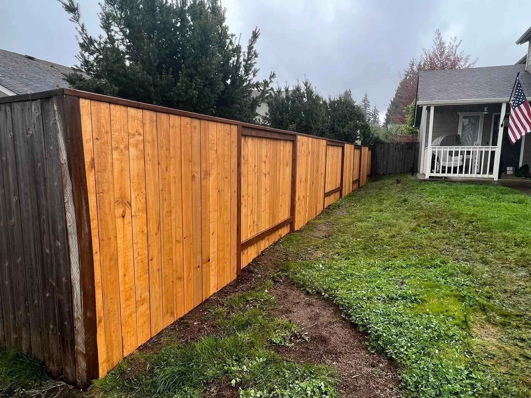 A backyard scene showing a newly installed wooden fence on the left side, and an older, darker fence on the far side. The yard has a patch of grass and some exposed soil. On the right side, there's a house with a small porch, an American flag hanging