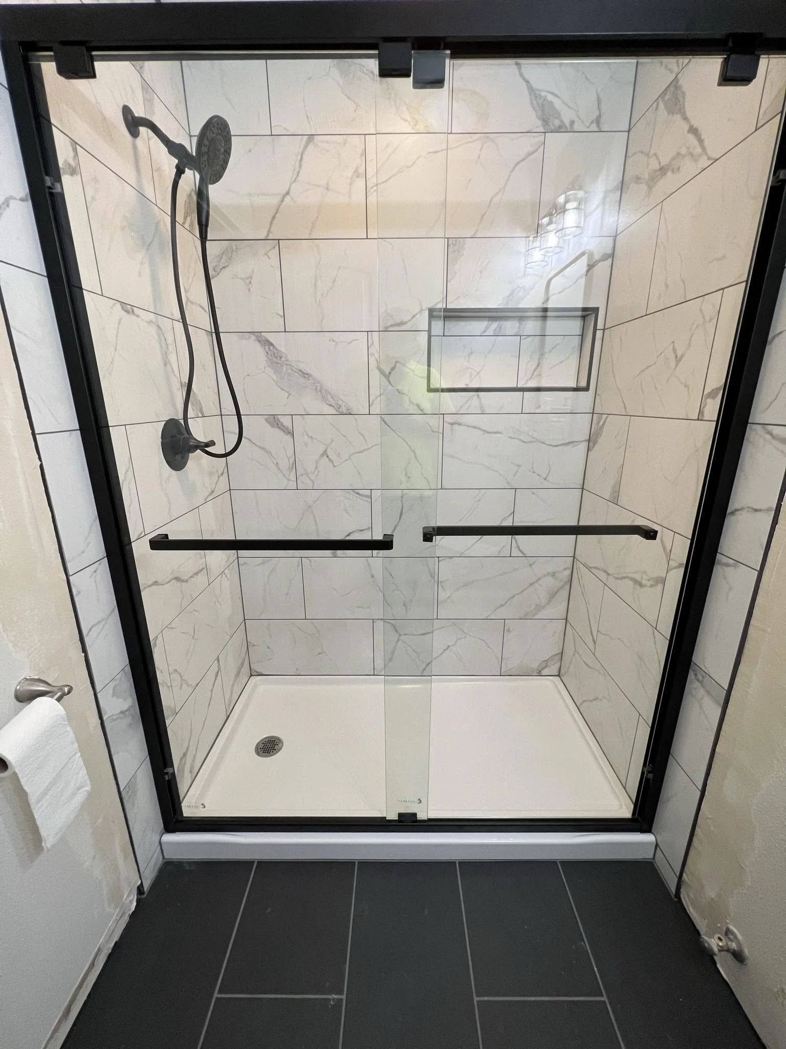 A glass shower enclosure with black framing, white marble-patterned tiles, a built-in niche, and dark floor tiles.