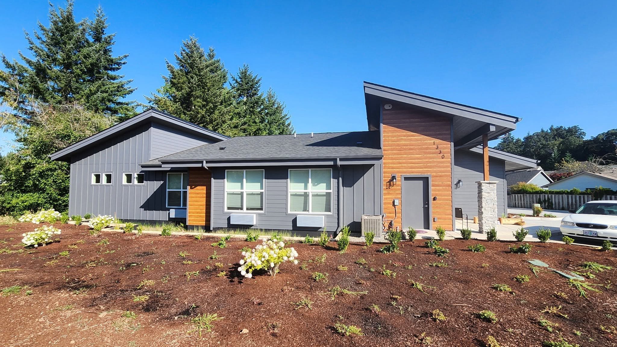 Modern house with gray and wood exterior, large windows, landscaped front yard under a clear blue sky.