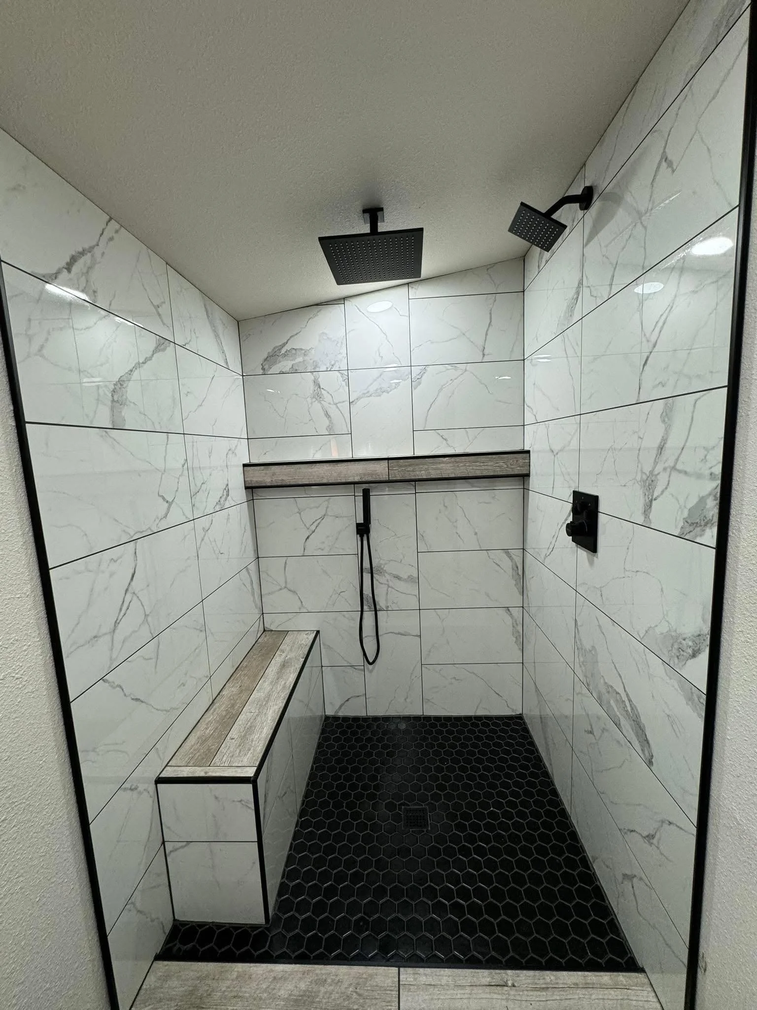 Modern bathroom shower with marble tiles, black rainfall showerhead, handheld shower, built-in bench, and black hexagonal floor tiles.