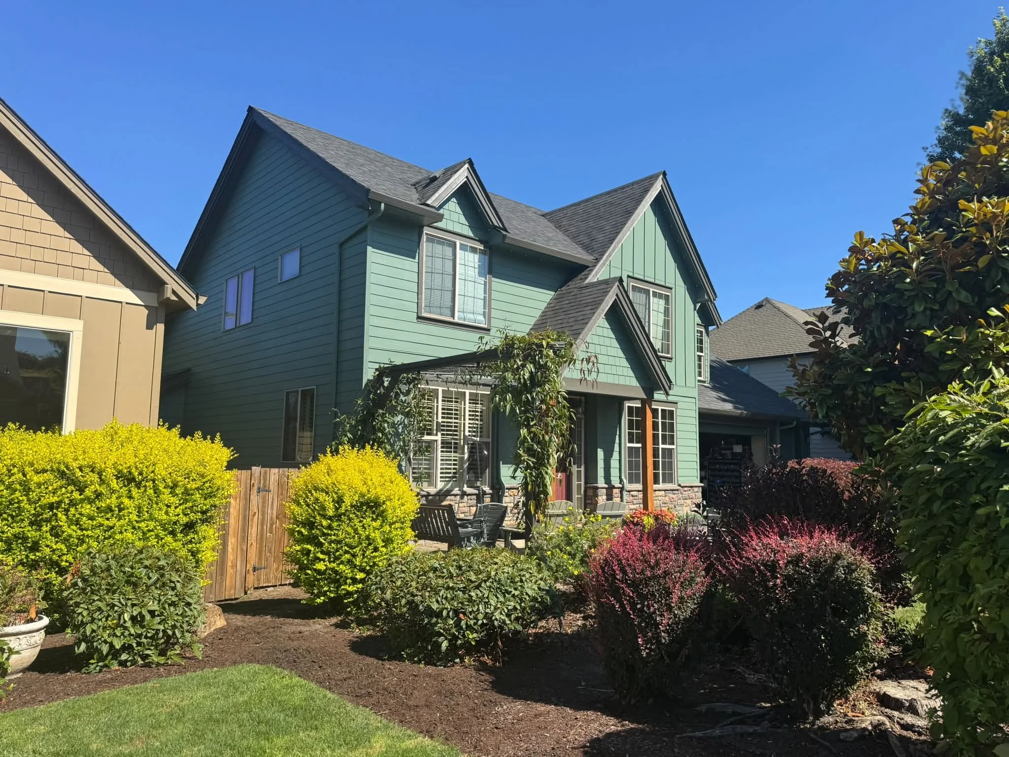 Colorful landscaped backyard with green lawn, flower bushes, shrubs, and trees, with a house featuring green siding and large windows under a clear blue sky.
