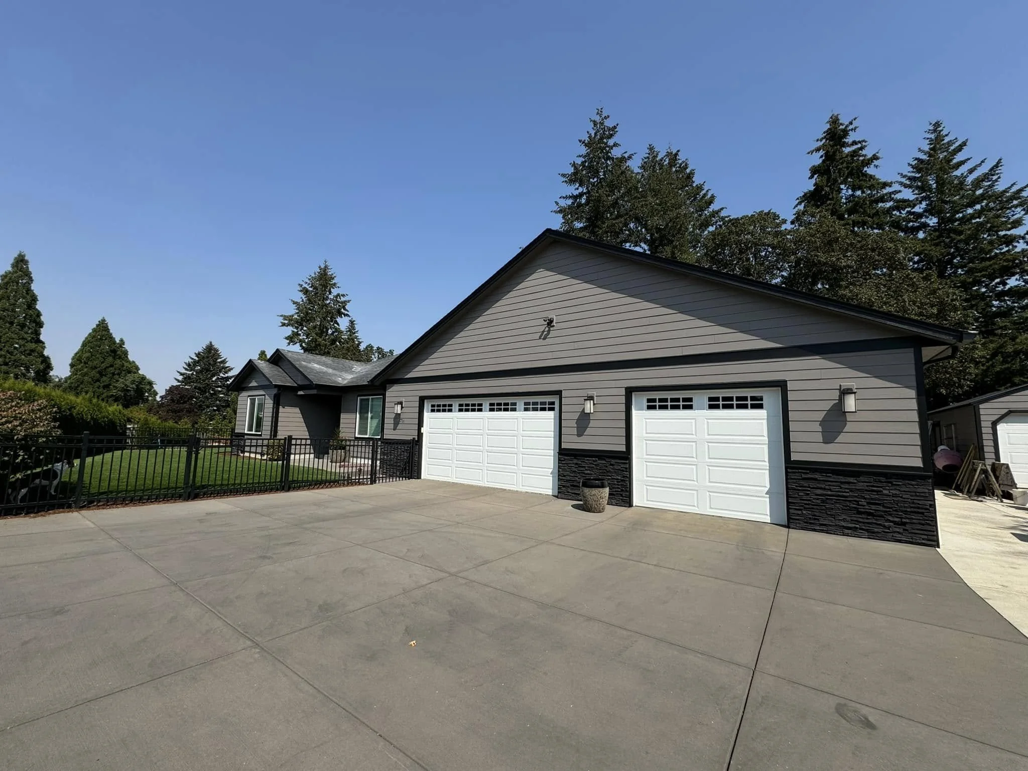 A house with a gray exterior, white garage doors, and black fence, situated on a large concrete driveway with green grass and trees in the background, under a clear blue sky.