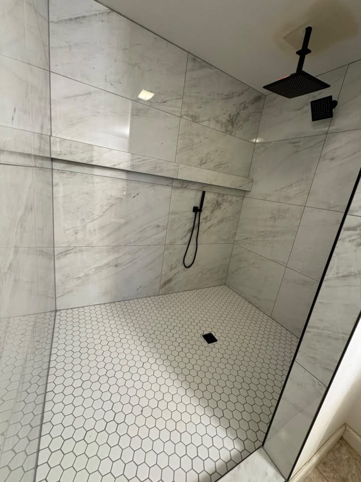Minimalist walk-in shower with large marble tiles, a black showerhead, a black handheld shower, and hexagon white floor tiles.