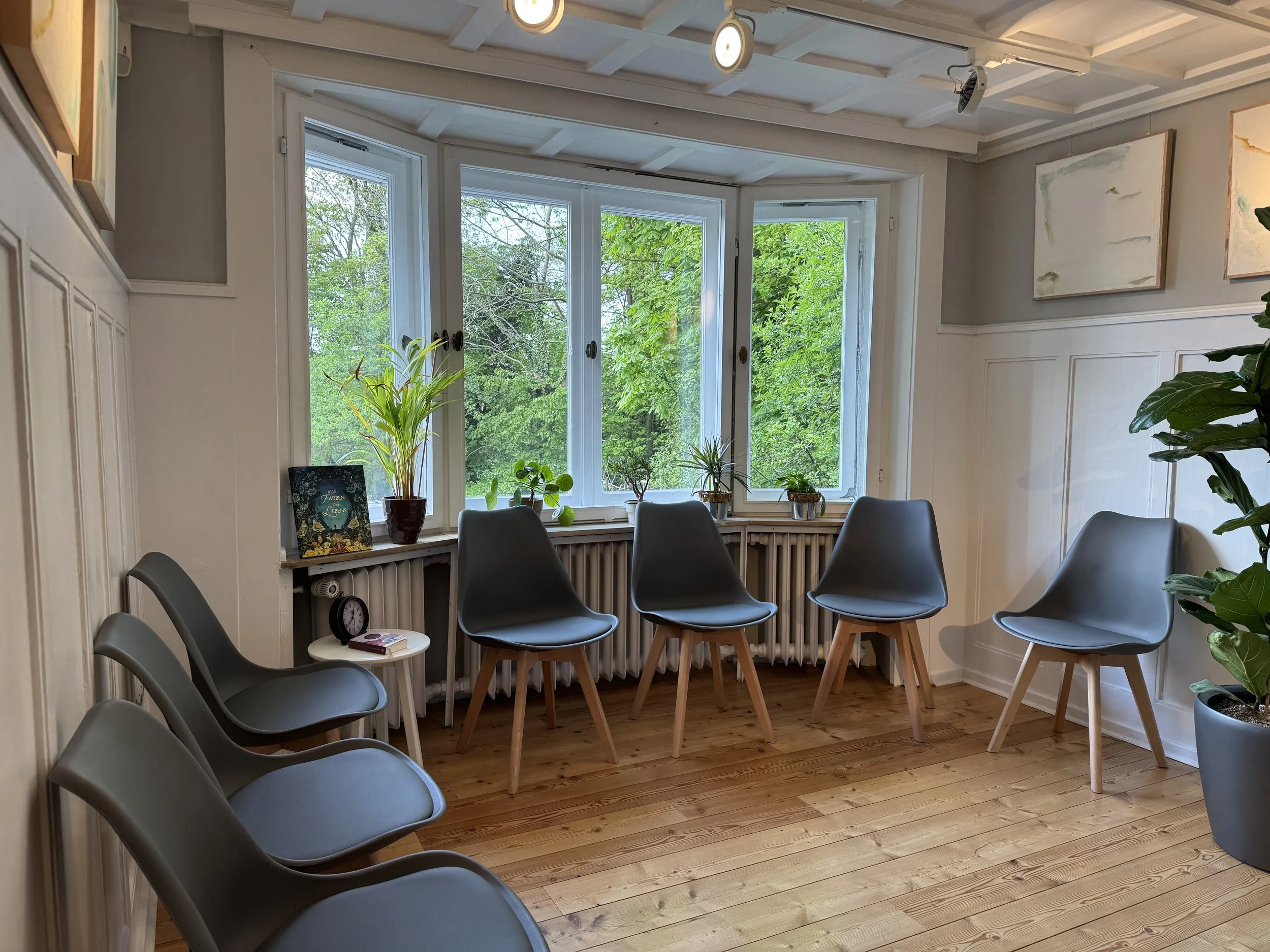 Innenraum eines Raumes mit mehreren Stühlen, große Fenster mit Blick auf grüne Bäume, einige Pflanzen auf der Fensterbank, Holzfußboden, Kunstwerke an der Wand, Uhren auf einem kleinen Tisch.