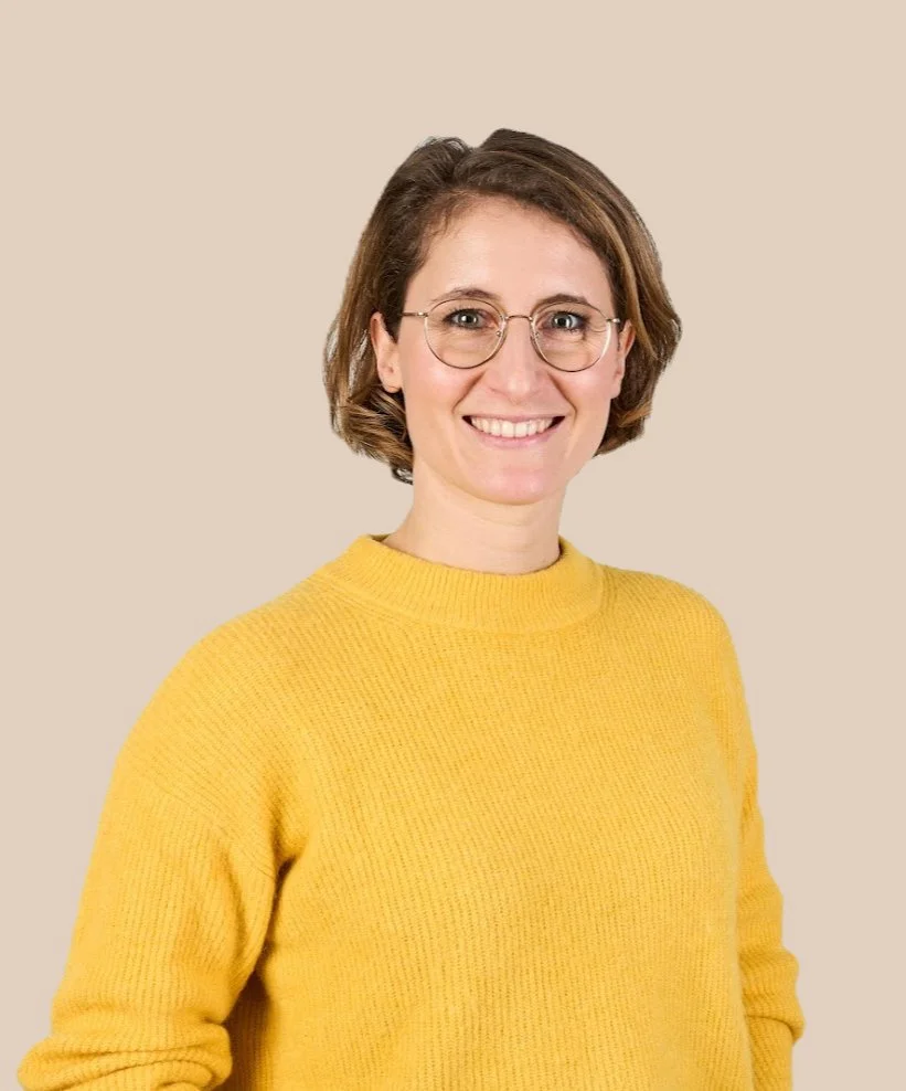 Eine Frau mit Brille, braunen kurzhaarigen Haaren und gelbem Pullover, smiling, vor neutralem Hintergrund.