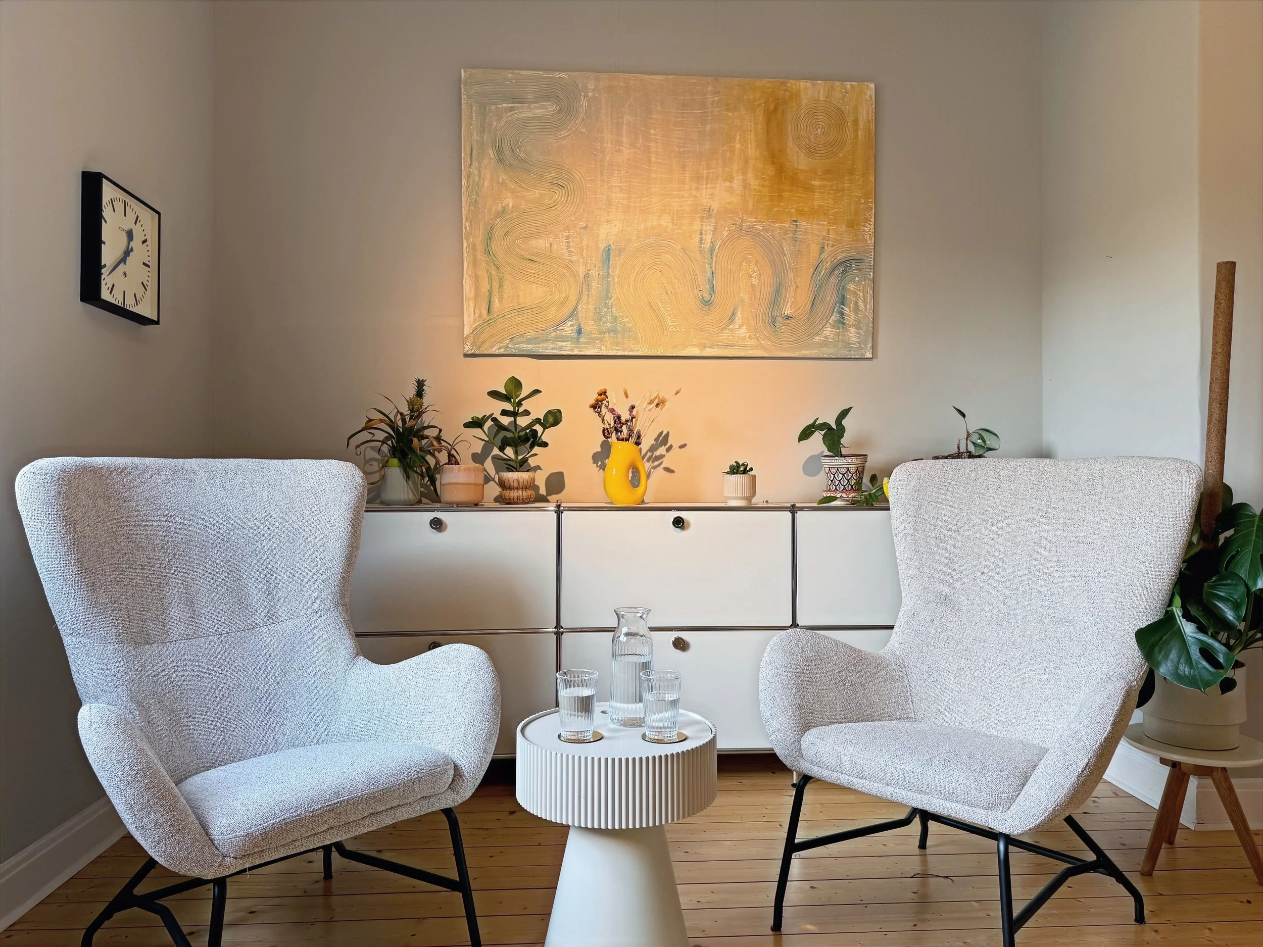 Ein Wohnzimmer mit zwei weißen, gepolsterten Sesseln um einen weißen, runden Beistelltisch, auf dem vier Gläser Wasser stehen. Im Hintergrund eine weiße Kommode mit mehreren Pflanzen, einer gelben Vase und einem Kunstwerk an der Wand. An der linken Wand eine Uhr. Auf dem im Hintergrund sichtbaren rechteckigen Kunstwerk sind geschwungene Linien und eine Wolke dargestellt.