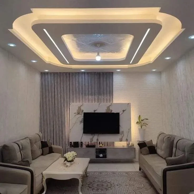 Modern living room with a decorative ceiling, wall-mounted TV on a marble accent wall, two beige sofas, a white coffee table with flowers, and a potted plant.