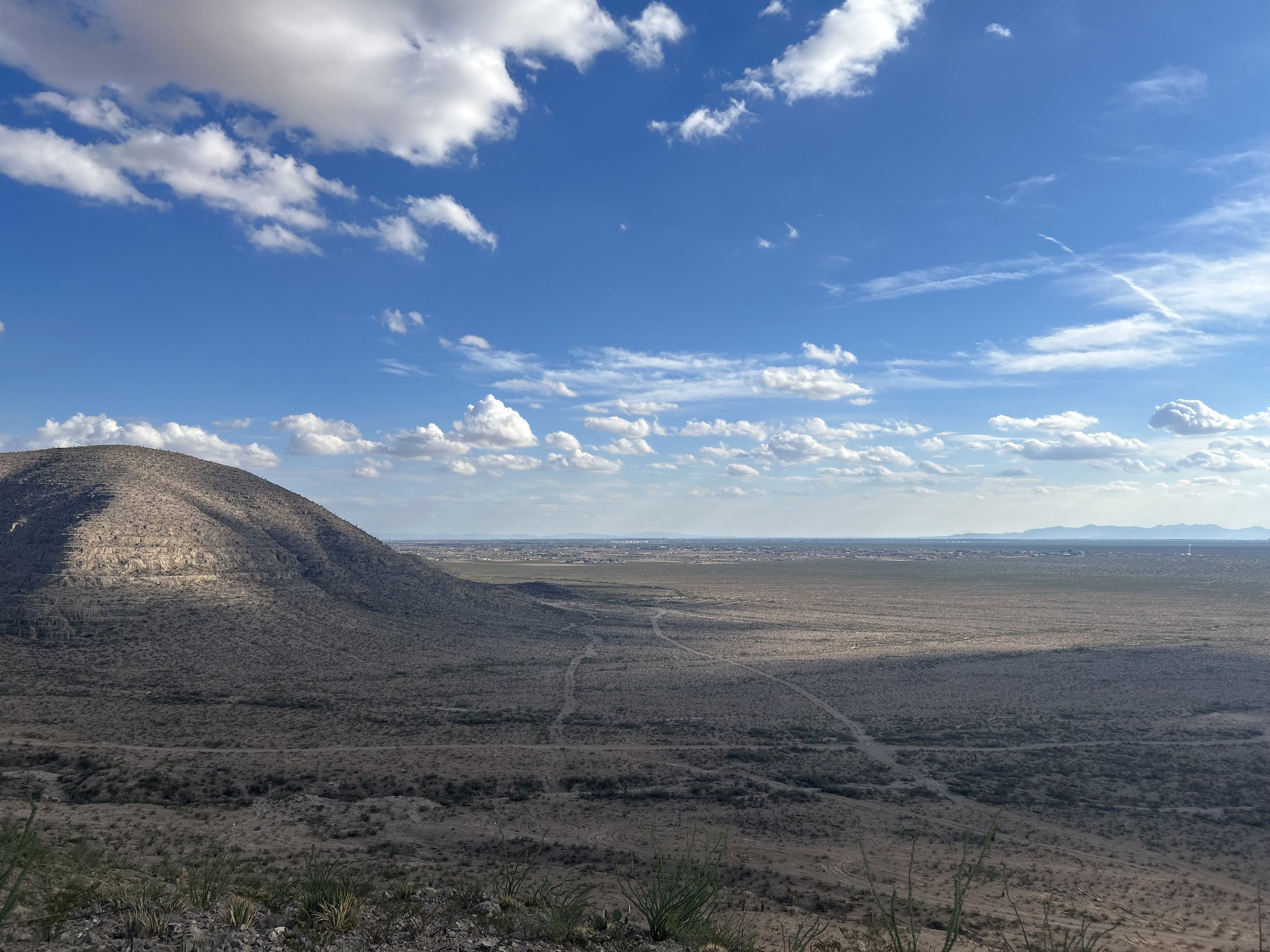 Open landscape with a large hill on the left, a flat plain extending to the horizon, and a blue sky with scattered white clouds.