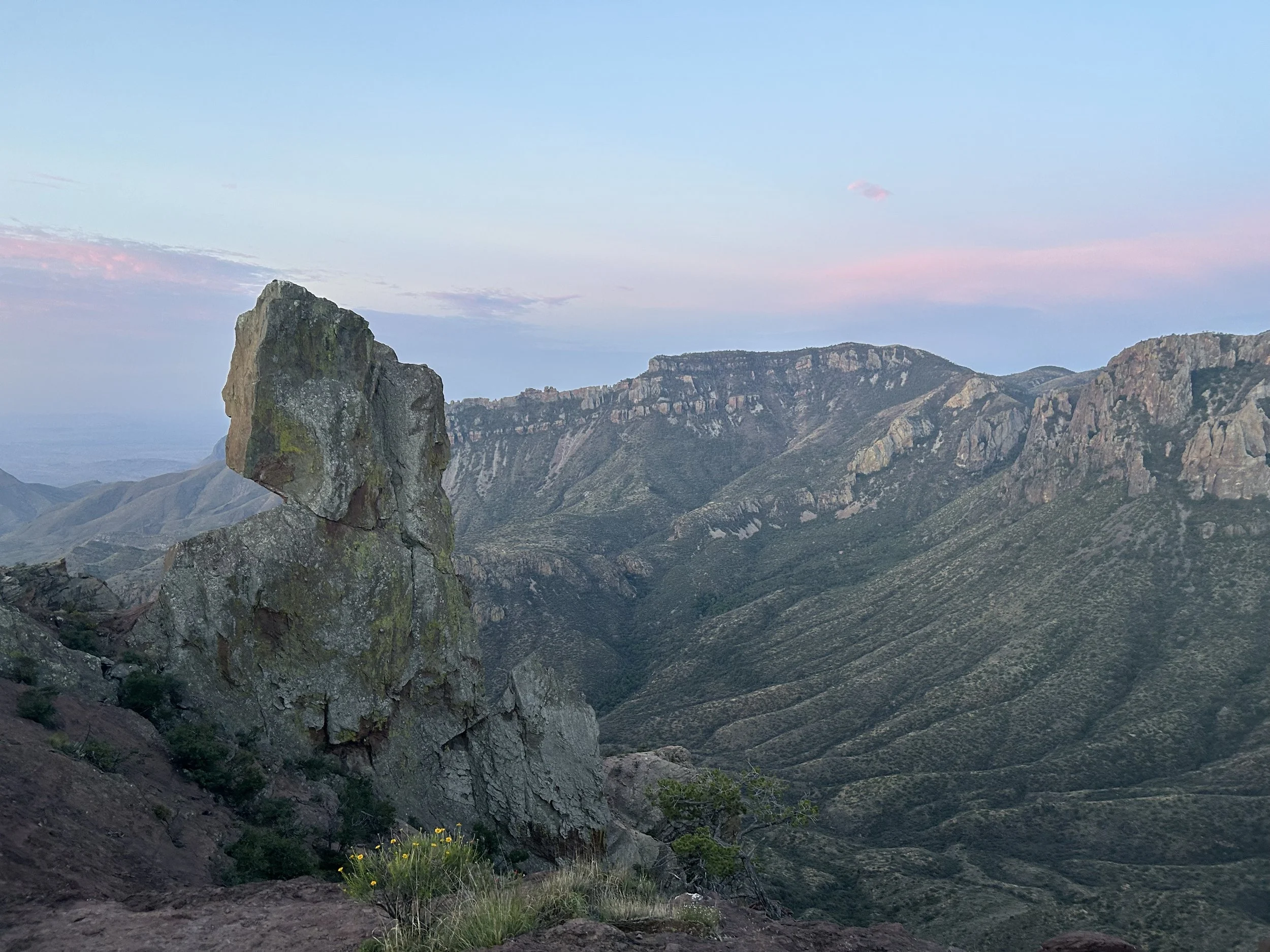 A rugged mountain landscape with a prominent large rock formation in the foreground, and a range of mountains with rocky cliffs and valleys in the background, under a sky with pastel-colored clouds.