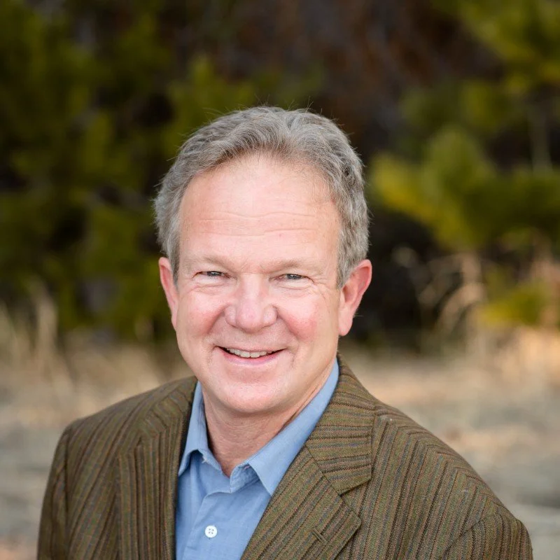 A smiling middle-aged man with short gray hair, wearing a brown blazer and light blue collared shirt, outdoors with blurred trees in the background.