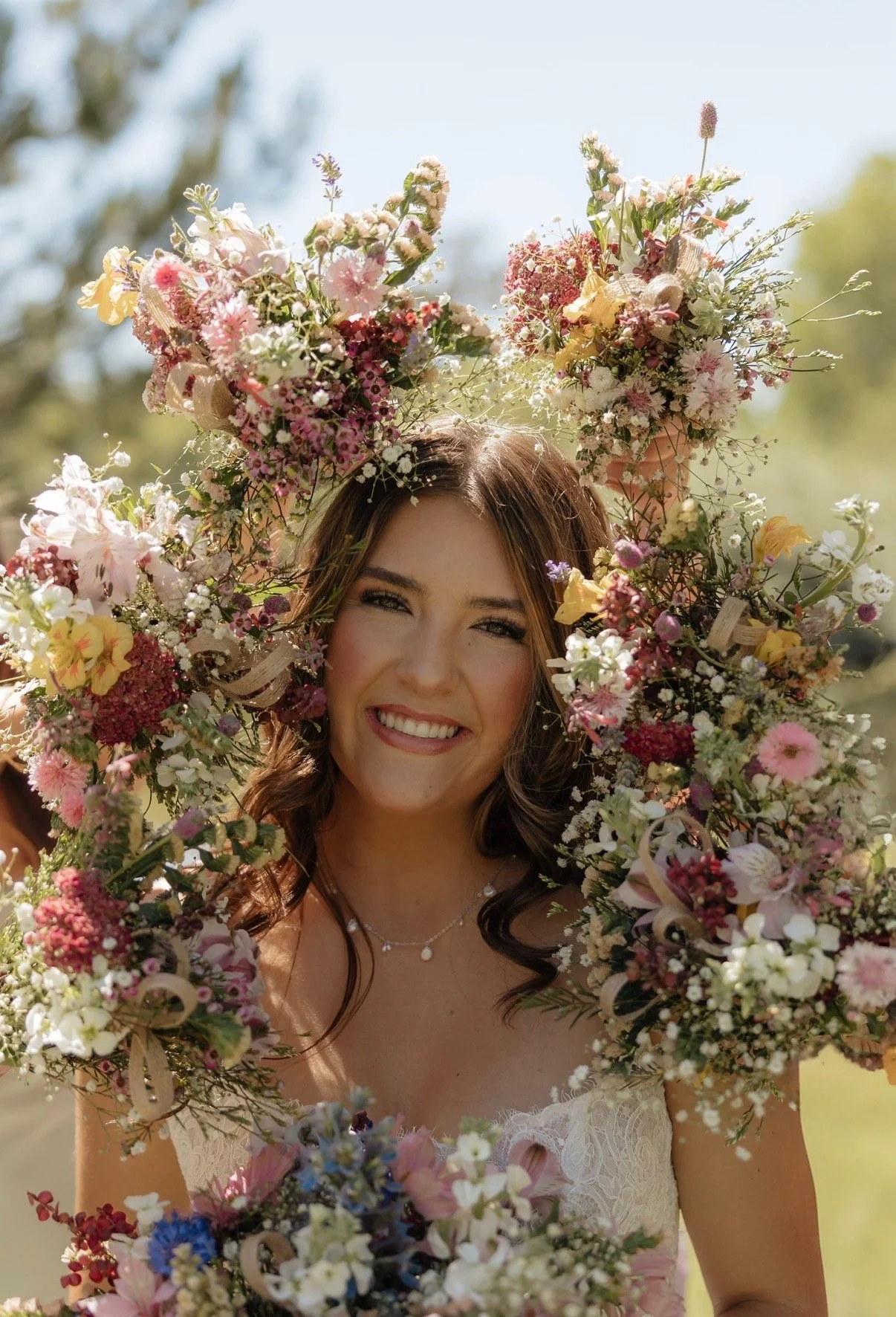 A smiling woman in a wedding dress holding a large floral arrangement shaped like a crown or arch, outdoors on a sunny day.