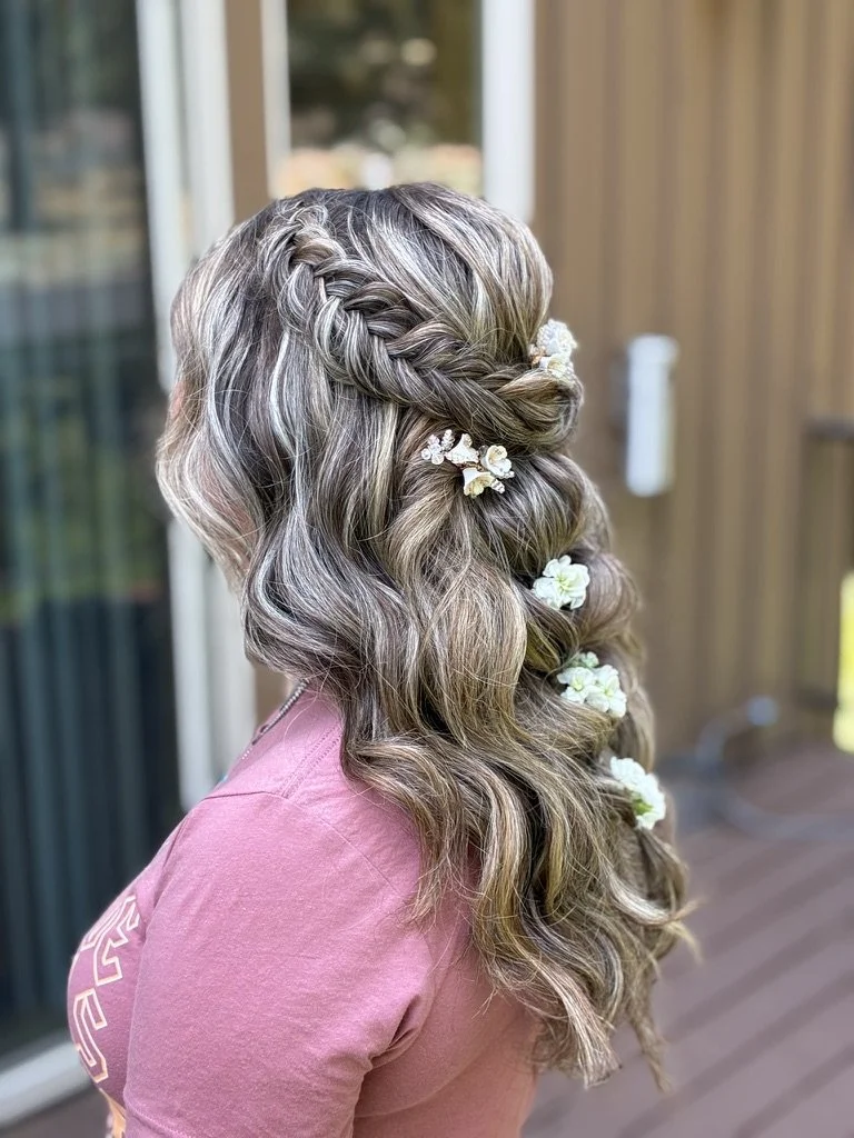 Woman with long, wavy, blonde hair styled with a braid and adorned with small white flowers, wearing a pink shirt, standing outdoors near a house.
