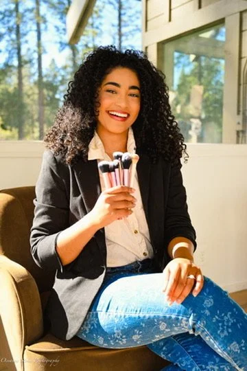 A woman sitting on a couch, smiling, holding a bundle of makeup brushes, in a bright room with large windows and trees outside.