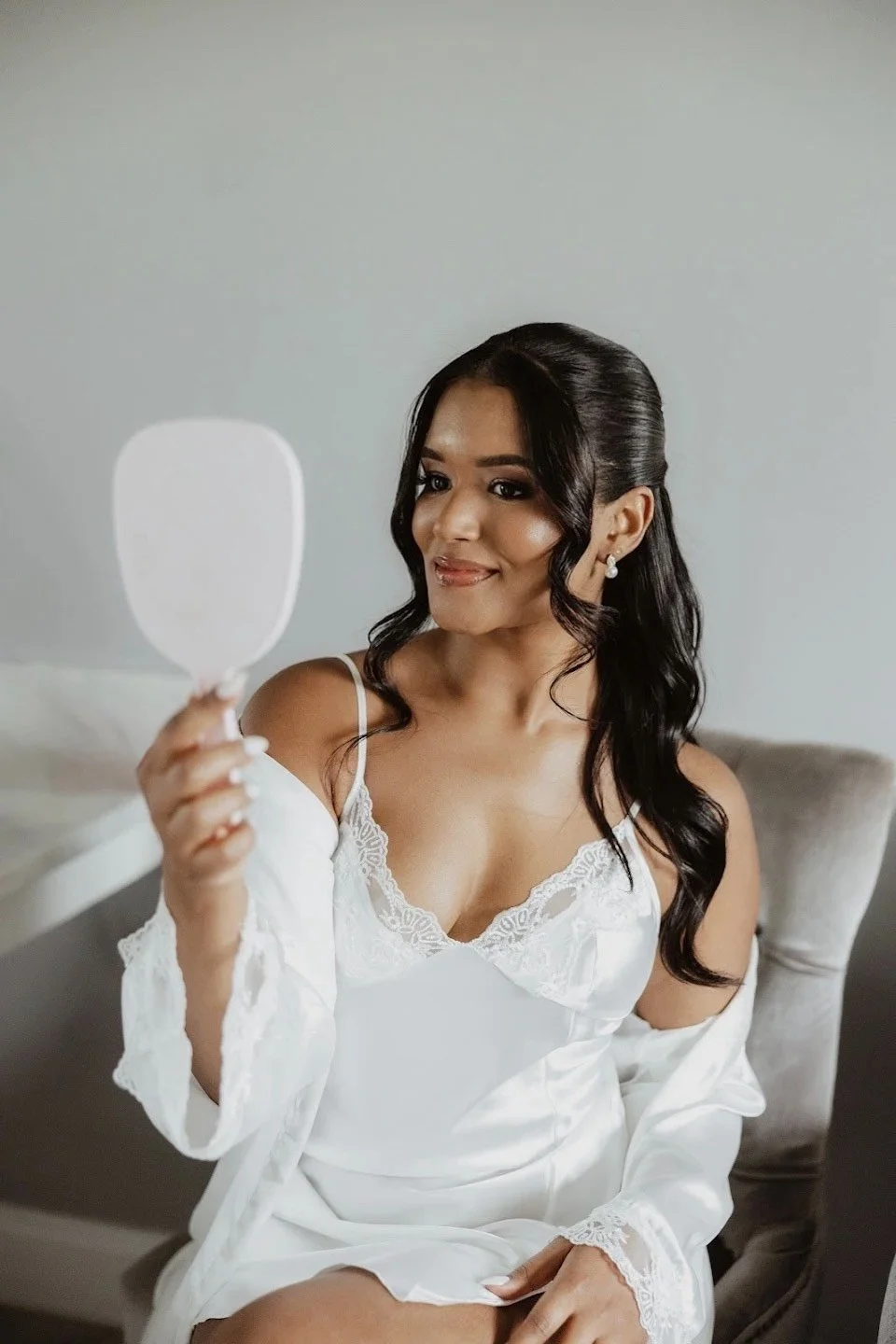 Woman wearing a white lace-trimmed silk satin nightgown, sitting on a chair, holding a hand mirror, in a bright room.