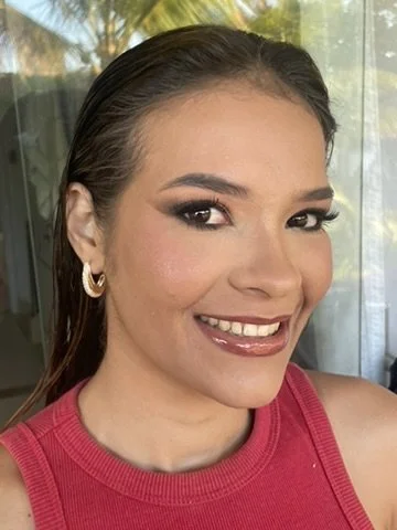 A woman with dark, wet hair, smiling, wearing gold hoop earrings and a pink sleeveless top, with natural light and greenery in the background.