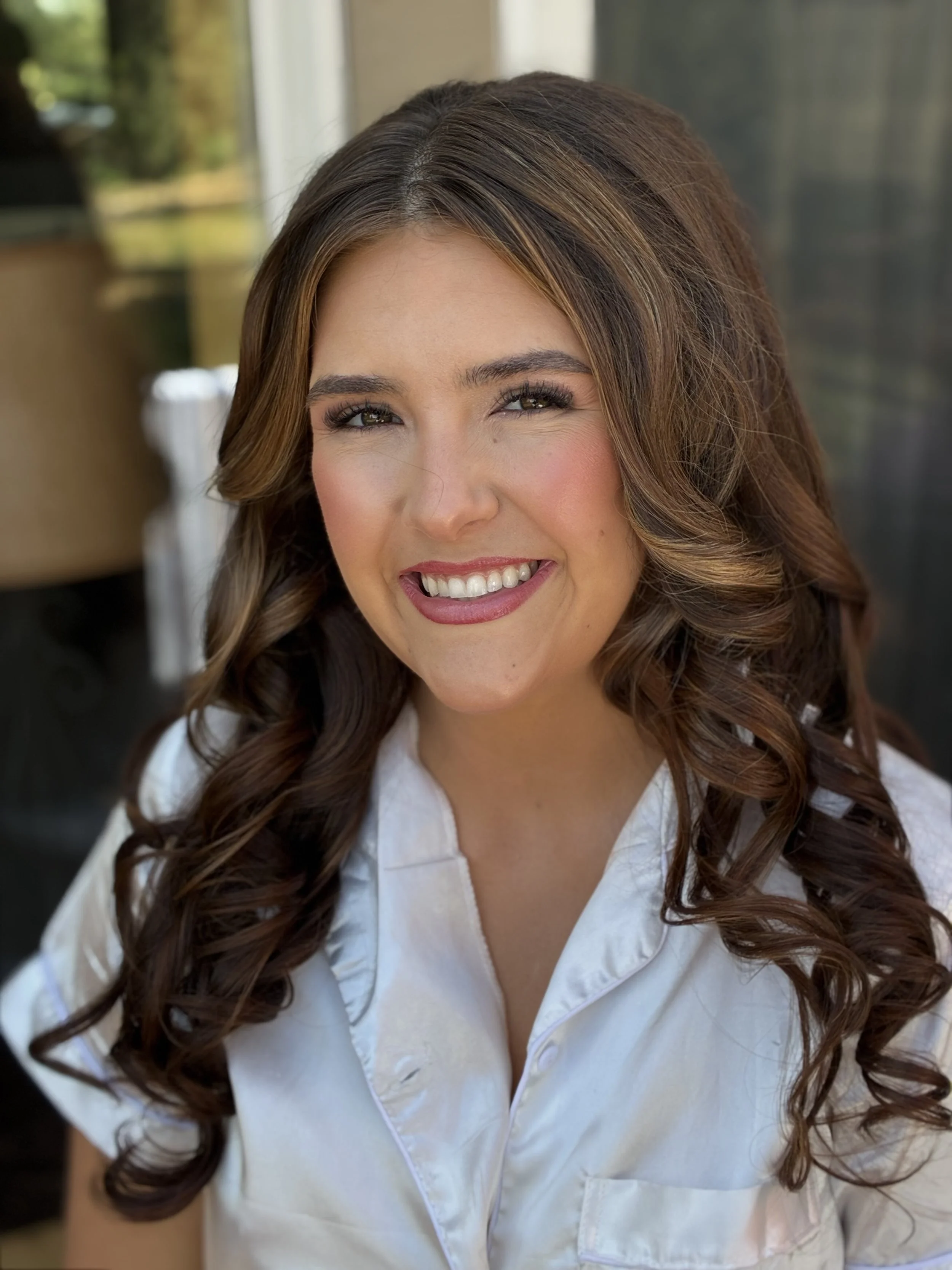 Close-up portrait of a woman with long brunette hair styled in loose curls, smiling with white teeth, wearing a white chef's coat.