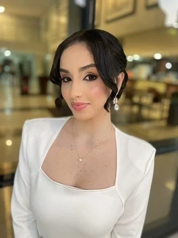 A woman with dark hair styled in loose waves, wearing a white dress with a square neckline and earrings, standing in a warmly lit indoor setting.