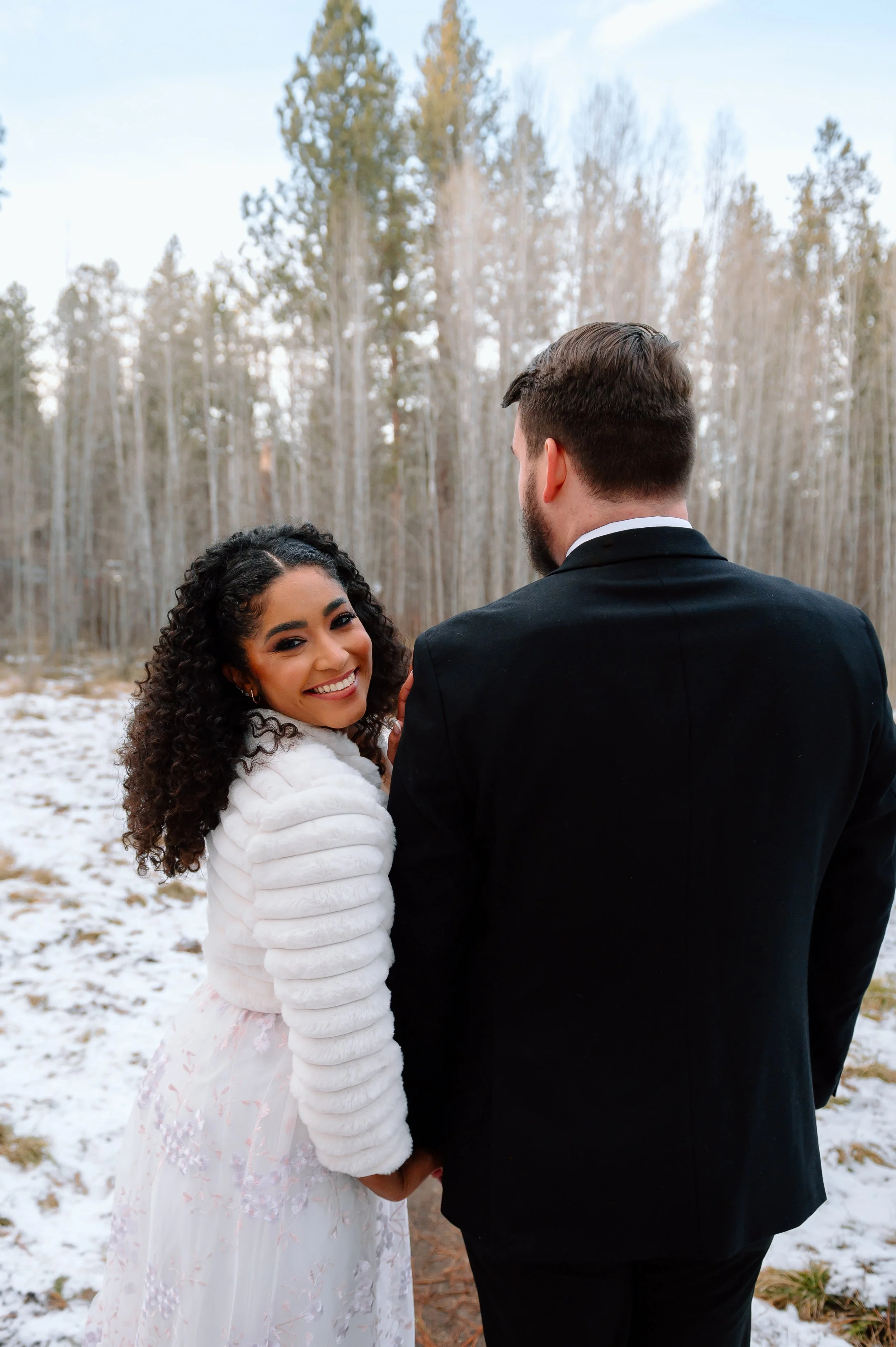 A smiling woman with curly hair in a white dress and fur coat holds hands with a man in a black suit outdoors in a snowy field with trees in the background.