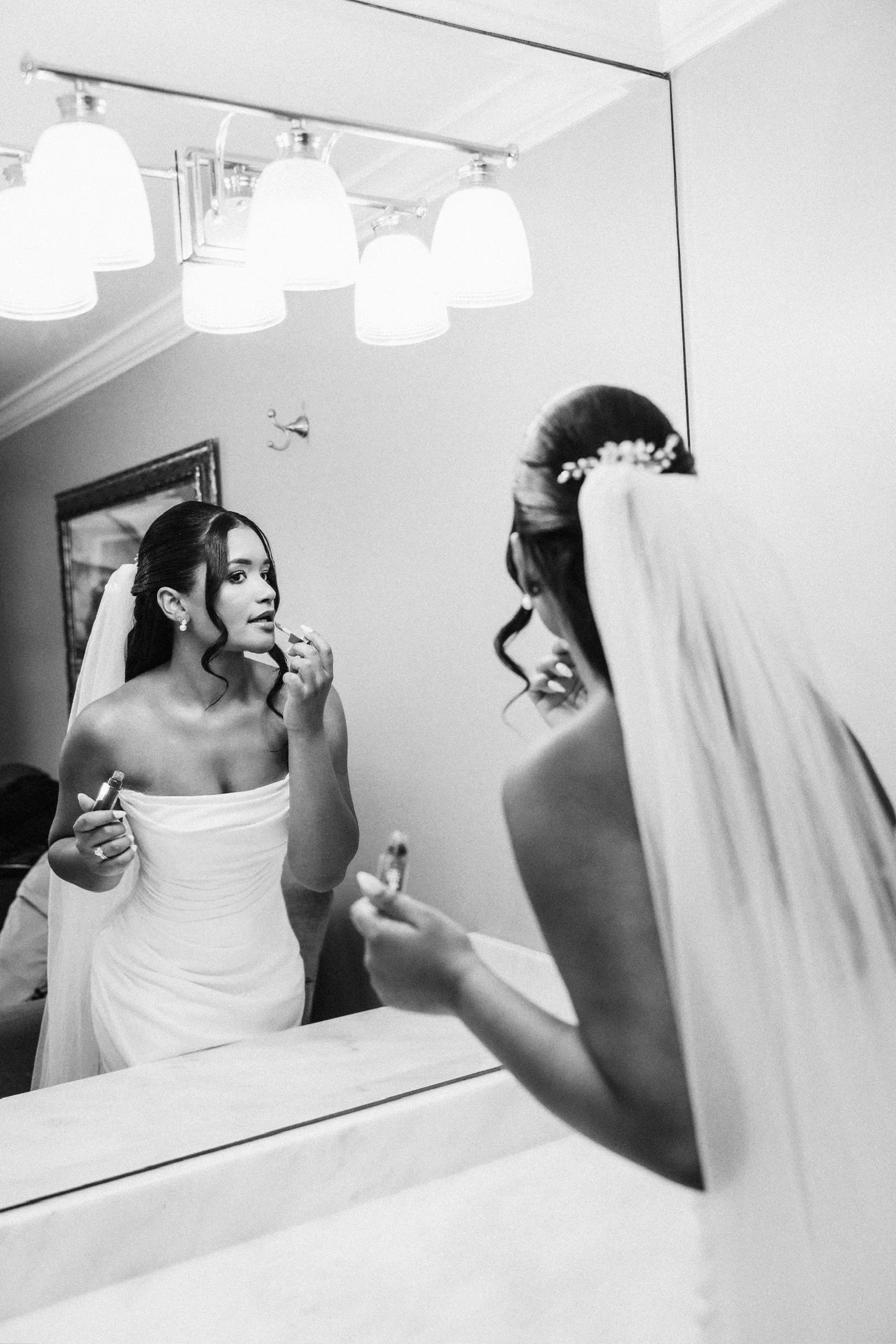 A woman in a white strapless dress is applying makeup while looking at her reflection in a mirror in a room with a ceiling light fixture.