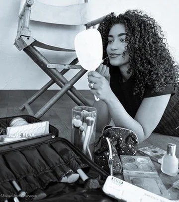 A woman with curly hair looking at herself in a mirror while sitting at a table surrounded by makeup and beauty products.