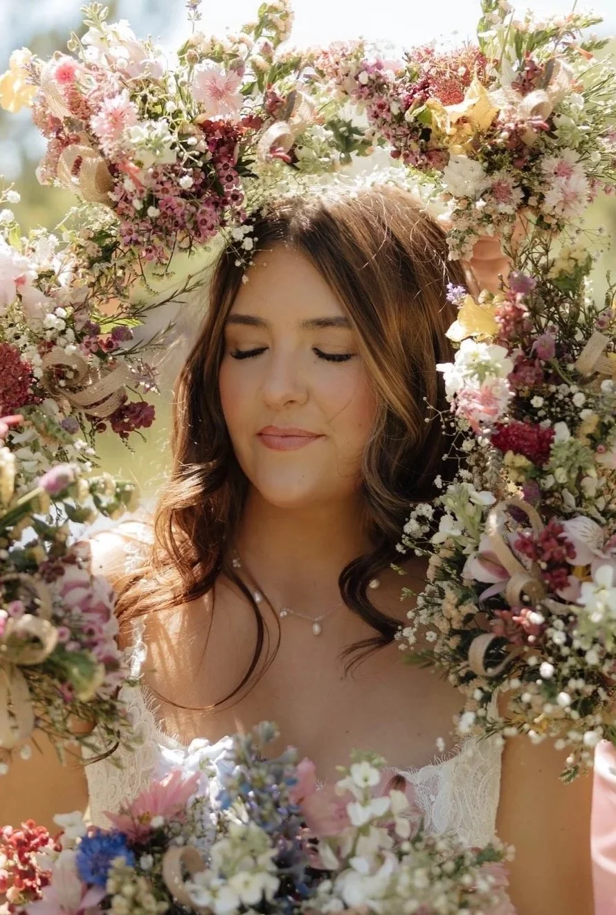 A young woman with closed eyes surrounded by a large, colorful floral wreath outdoors on a sunny day.