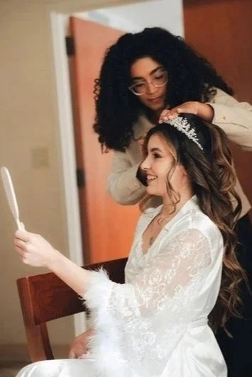 Two women, one with curly black hair and glasses, assisting the other with long, wavy hair and a tiara, in a room with a wooden door and beige walls. The woman with the tiara is dressed in a white lace robe and is holding a mirror, smiling as she looks at her reflection.
