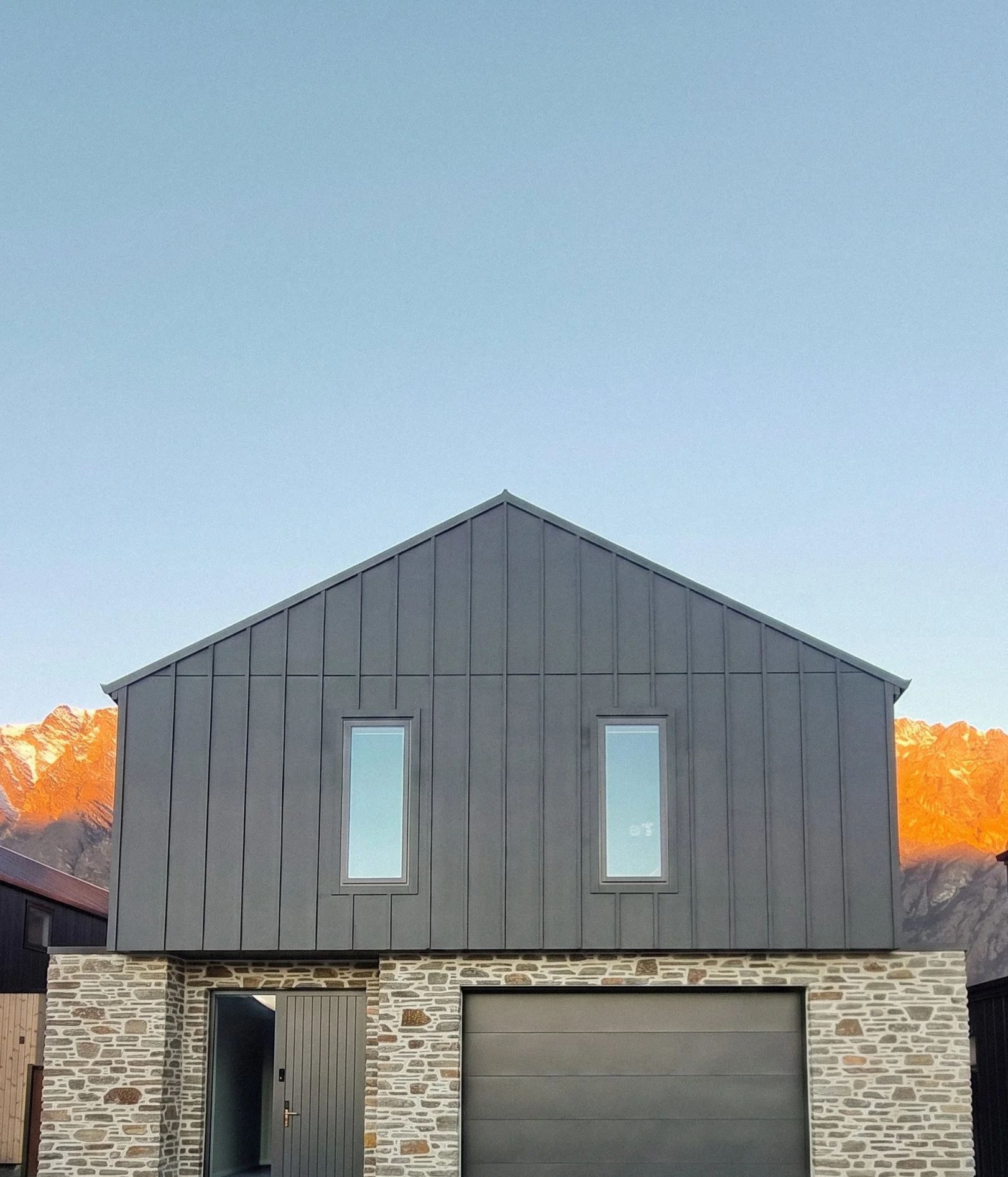 Modern house with Glenorchy Schist and James Hardie Board & Batten cladding, featuring a garage door, front door, and tall narrow windows, with The Remarkables Mountains in the background and a clear sky.