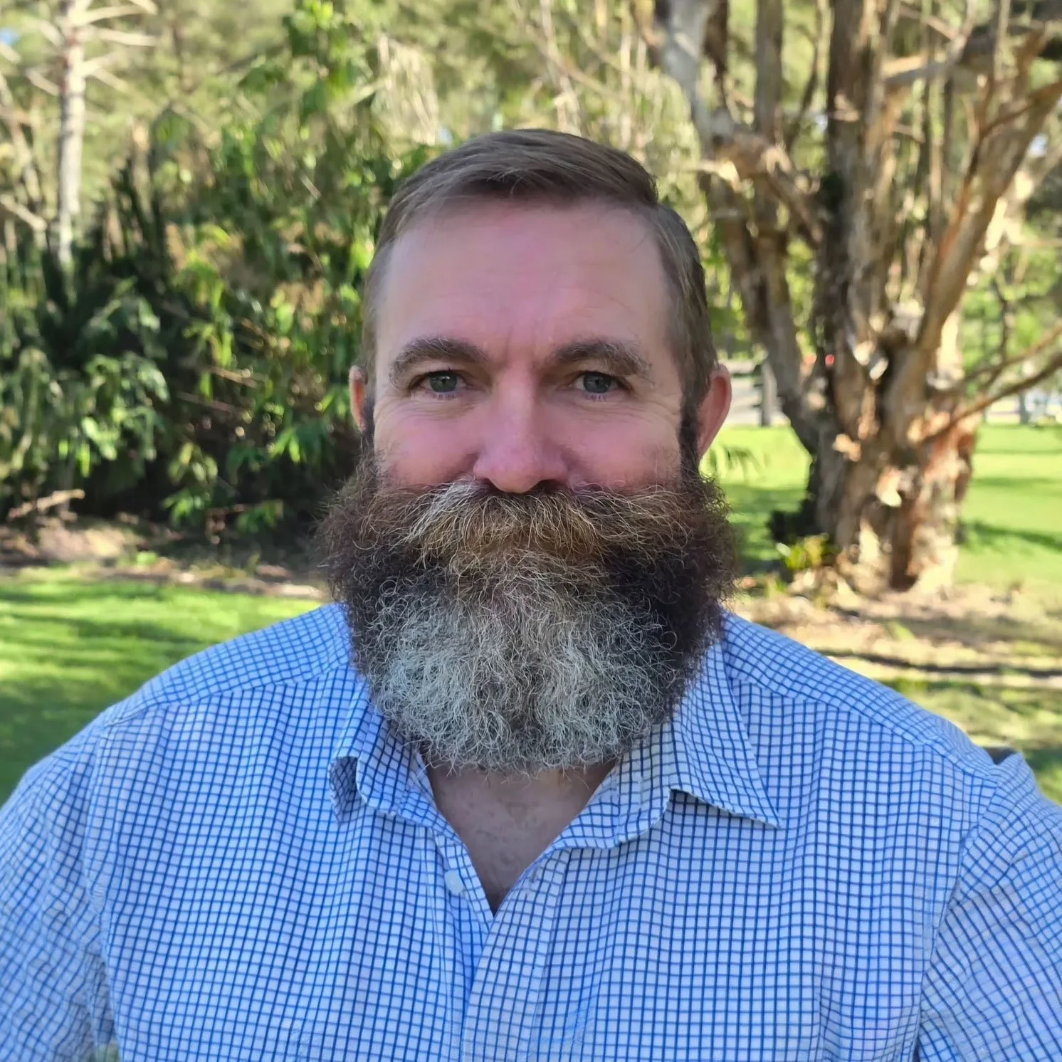 A man with a full beard and mustache wearing a blue checked shirt posing outdoors with trees and green grass in the background.