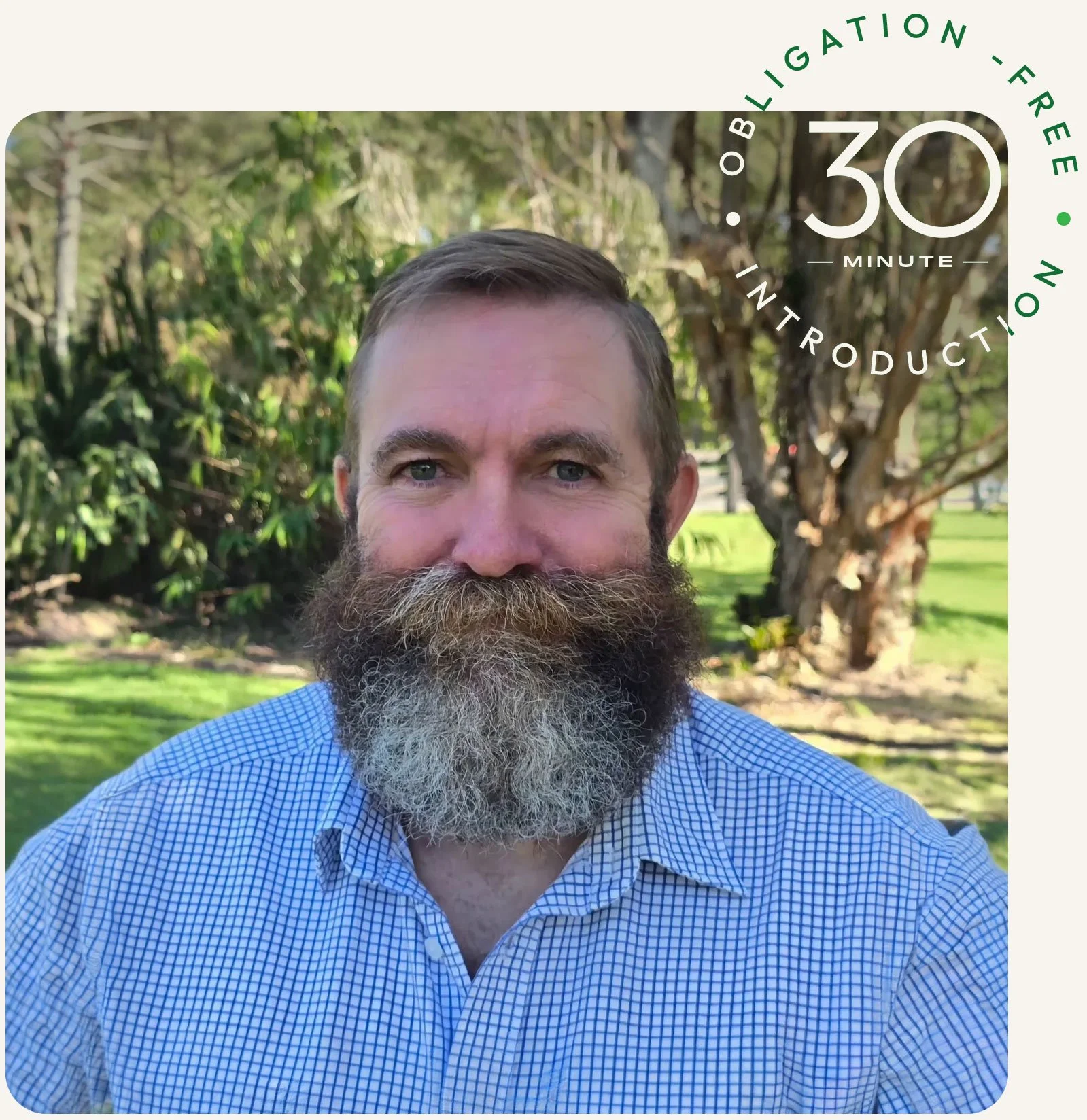A man with a beard and mustache wearing a blue checkered shirt taking a selfie outdoors with trees and grass in the background.