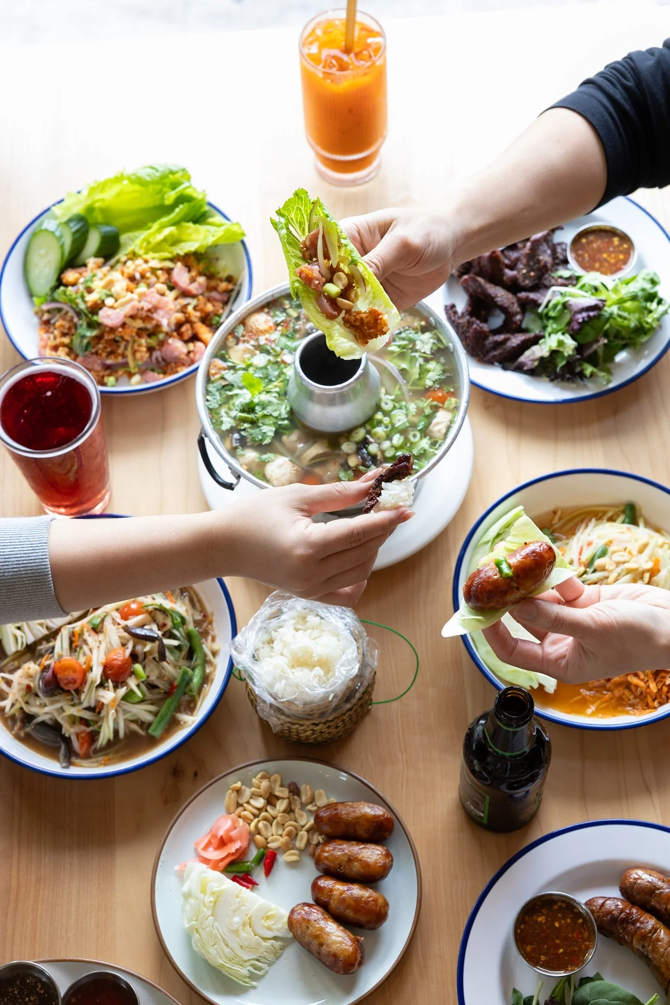 Various dishes of Asian cuisine, including spring rolls, salads, grilled meats, and dipping sauces, served on plates and bowls on a wooden table.