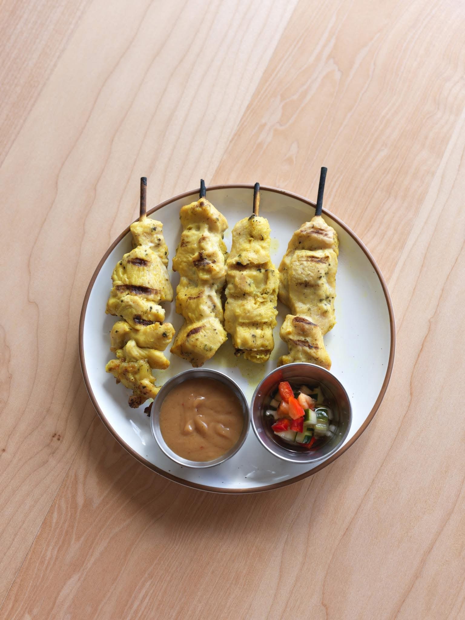 Plate with four grilled chicken skewers, two small bowls of dipping sauces, and a side of chopped vegetables.