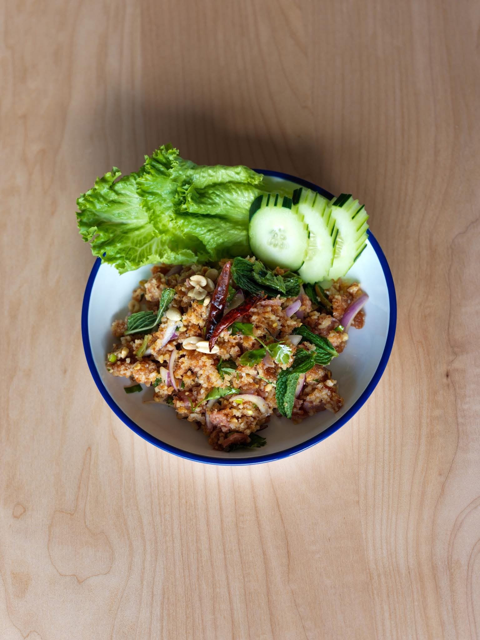 A bowl of Thai larb with chopped vegetables and herbs, garnished with slices of cucumber and lettuce on a wooden table.